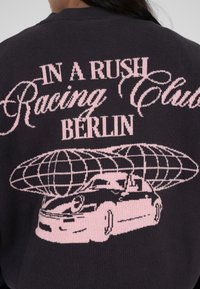 Pull en tricot noir avec texte et graphiques roses. Le texte inclut "IN A RUSH Racing Club BERLIN." Le design présente une voiture et un motif de globe.
