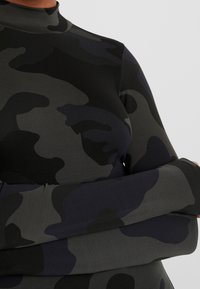 Camo-mönstrad långärmad topp i nyanser av grått och svart. Har en hög hals och en åtsittande design med slät, elastisk textur.