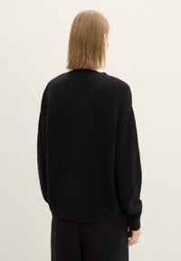 Musta neulottu villapaita, jossa on löysä istuvuus, resorikuviointi ja leveät hihat. Takana on hieman lyhyt helma ja pyöreä pääntie.