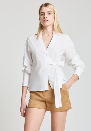 White button-up shirt with a tie detail at the waist; long sleeves with cuff; smooth, lightweight fabric; paired with tan shorts.