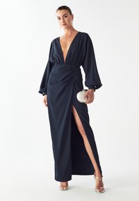 Námornícky dlhý šaty z hladkej tkaniny majú hlboký V-výstrih, zhrnutý pás, dlhé rukávy, bočný rozparok a elegantnú striebornú kabelku.