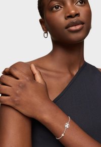Donna che indossa orecchini a cerchio in argento e un braccialetto d'argento con un charm a forma di globo circolare e una perla, vestita con un top senza maniche nero.