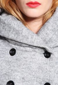Närbild på en person med röd läppstift som bär en strukturerad grå kavaj med stora svarta knappar och en bred krage.