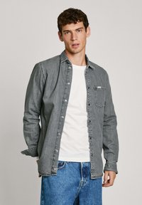 Grijze denim overhemd met een knoopsluiting aan de voorkant, één borstzak en lange mouwen, gedragen over een wit t-shirt en blauwe jeans.