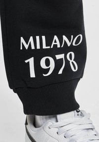 Schwarze Jogginghosen mit weißem Aufdruck "MILANO 1978". Der Stoff ist glatt mit gerippten Bündchen; kombiniert mit weißen Sneakers.