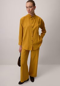 Hořčicově žlutý set košile a kalhot, který má zap�ínání na knoflíky, dlouhé rukávy a jemné ozdoby. Doplněno tmavohnědými doplňky.