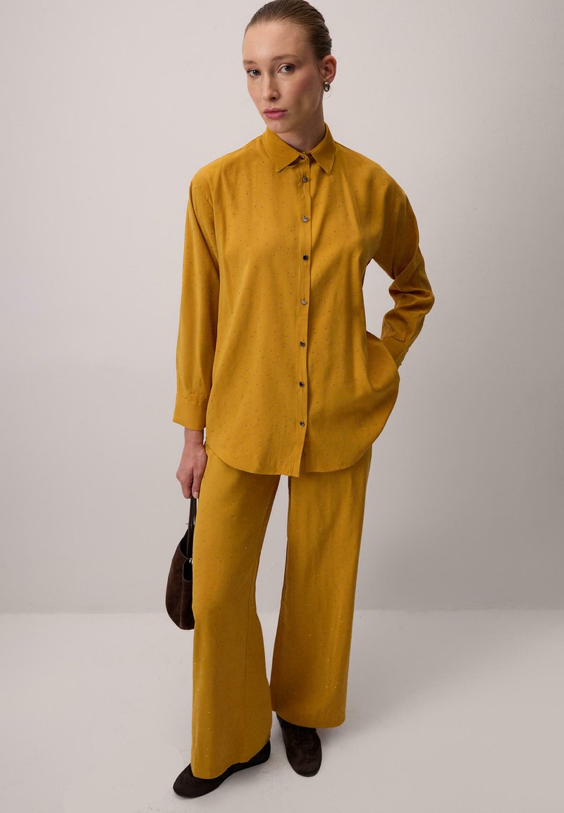Hořčicově žlutý set košile a kalhot, který má zapínání na knoflíky, dlouhé rukávy a jemné ozdoby. Doplněno tmavohnědými doplňky.