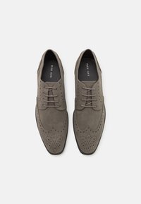 Une paire de chaussures brogue pour homme en suède gris avec des détails en pointe perfor ée, des semelles noires et des lacets sur fond blanc.