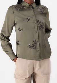 Blusa de satén verde oliva con botones al frente, mangas largas y adornos de lentejuelas florales en el pecho y los brazos. Combinada con pantalones beige.