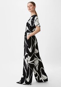 Czarny i biały kombinezon z abstrakcyjnymi wzorami, z krótkimi rękawami, szerokimi nogawkami i dopasowaną talią. Posiada boczne kieszenie oraz pasek do wiązania.