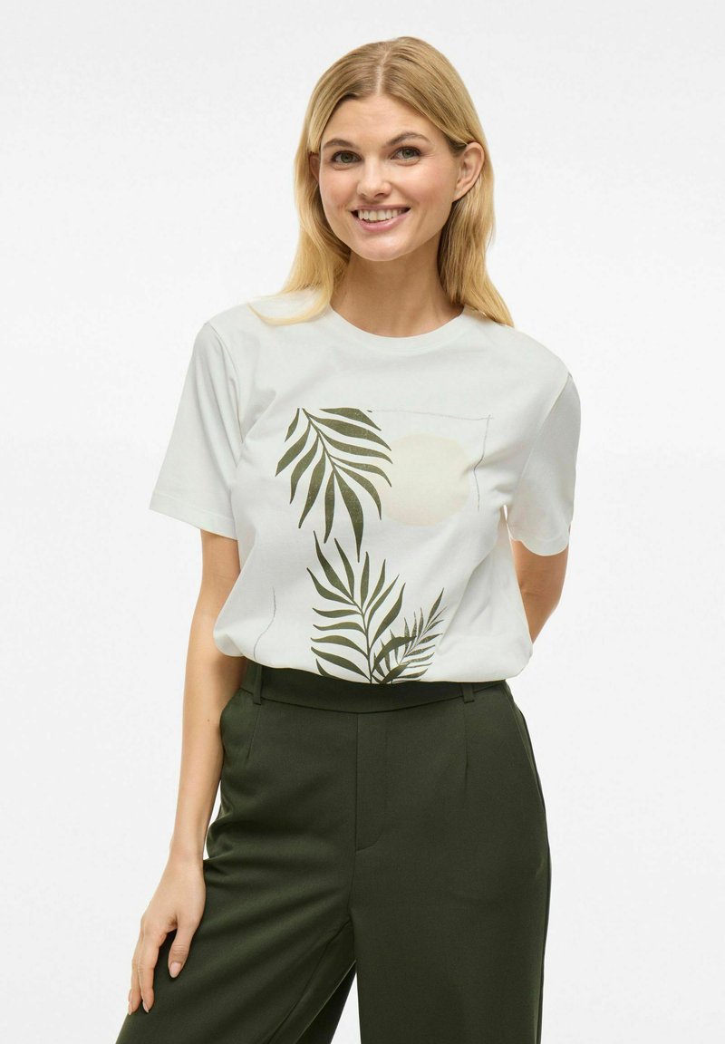 Valkoinen lyhythihainen t-paita, jossa on vihreä lehtikuvio ja pyöreä muotoilu. Yhdistetty tummanvihreisiin korkeavyötäröisiin housuihin. Silkkinen pinta.