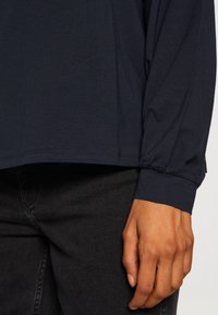 Navy long-sleeve shirt with gathered cuffs, made from soft fabric, paired with dark jeans visible at the bottom.