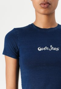 Sötétkék, testhezálló póló, puha anyagból készült, rövid ujjú, kerek nyakkal, az elején fehér "Guess Jeans" logóval.