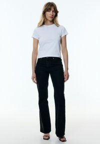 Weißes Baumwoll-T-Shirt mit kurzen Ärmeln, kombiniert mit schwarzer, hochtaillierter, ausgestellter Jeanshose und schwarzen, hohen Sandalen. Einfaches, figurschmeichelndes Design.