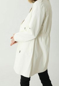 Chaqueta beige claro con cintura ceñida, mangas largas y bolsillos laterales. El material parece suave con un ligero brillo. Diseño de cuello sencillo.