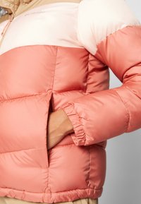 Ręka w kieszeni różowo-kremowej pikowanej kurtki z elastycznymi mankietami i widocznym logo Columbia przy ramieniu.