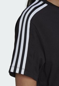 Close-up van een persoon die een zwart T-shirt met korte mouwen draagt, waarop drie witte strepen naar beneden over de schouder en mouw lopen.