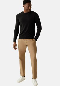 Svart långärmad tröja, beige byxor med en slät textur och vita sneakers. Modellen står med händerna i fickorna och ler.