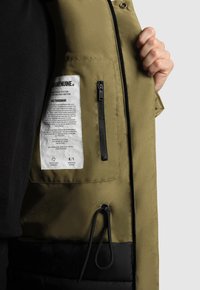 Olīvu zaļas jakas iekšpuse ar auduma kopšanas etiķeti, melnu rāvējslēdzēju kabatu un auklu. Gluda, matēta tekstūra.