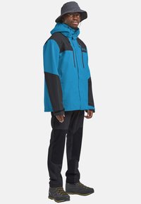Veste imperméable bleue et noire avec capuche réglable, poches zippées, pantalon noir assorti et bottes grises avec accents jaunes.