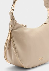Beige Lederhandtasche mit goldenen Reißverschluss und Logo, runder Form und verstellbarem Trageriemen, vor einem hellen Hintergrund.