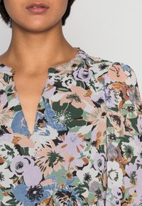 Blouse florale avec un col en V, des manches courtes bouffantes et un mélange coloré de fleurs vertes, roses et bleues sur un fond clair.