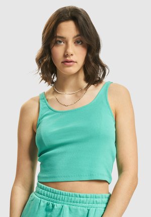 Young woman with wavy brown hair wearing a turquoise cropped tank top and matching sweatpants against a plain background.