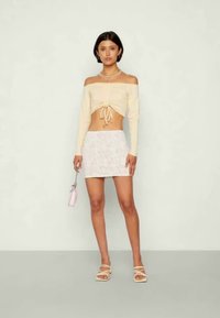 Top crop bege claro, com ombros à mostra e cordão, combinado com uma saia mini branca com padrão floral. Acessorizado com sandálias de tiras e uma bolsa rosa.
