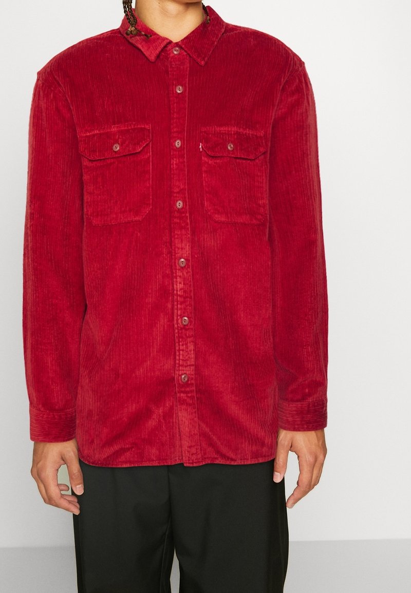 Camisa de pana roja con cierre de botones, dos bolsillos en el pecho y cuello clásico. Tejido texturizado con rayas verticales, mangas largas.