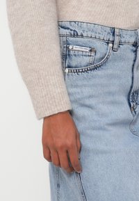 En venstre hånd på en person iført en beige strikket sweater hviler ved siden af lys blå denimbukser med en synlig lille lomme og et mærke.