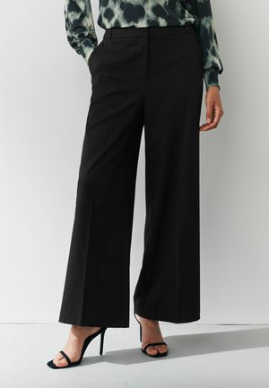 Schwarze weite Hosen mit einem glatten, matten Finish und seitlichen Taschen, kombiniert mit schwarzen Sandalen mit Riemchen und hohen Absätzen.