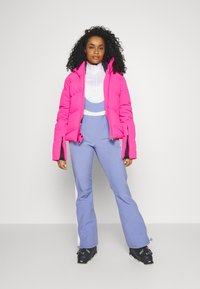 Heldere roze pufferjack met ritsmanchetten, witte basislaag en blauwe skibroek met witte zijaccenten. Zwarte skimenner maken de look compleet.