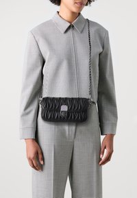 Czarna pikowana torebka na ramię z łańcuszkiem, o prostokątnym kształcie i metalicznym zapięciu. Nosi ją osoba w kostiumie w kratkę.