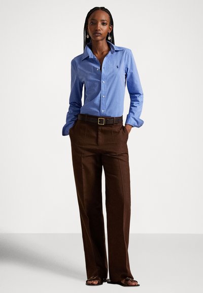 Ανοιχτό μπλε πουκάμισο με κουμπιά και γιακά, συνδυασμένο με καφέ, ραμμένο παντελόνι. Το πουκάμισο φέρει ένα λογότυπο και μια μαύρη ζώνη τονίζει τη μέση.