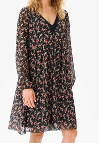 Nainen, jolla on päällään musta läpikuultava pitkähihaisellinen mekko, jossa on pieniä vaaleanpunaisia, valkoisia ja punaisia kukkakuvioita, sekä V-kaula-aukollinen musta aluspaita.