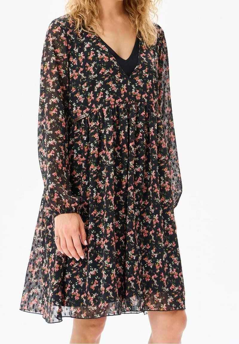 Nainen, jolla on päällään musta läpikuultava pitkähihaisellinen mekko, jossa on pieniä vaaleanpunaisia, valkoisia ja punaisia kukkakuvioita, sekä V-kaula-aukollinen musta aluspaita.