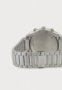 Silberne Metall-Armbanduhr mit einem gemusterten Gliederarmband, ausgestattet mit einem runden Gehäuse, grünen Akzenten an der Krone und einer polierten Oberfläche.