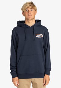 Sudadera azul marino con un bolsillo de canguro en la parte delantera, capucha con cordón y un pequeño parche con el logo de Billabong en el pecho. Fabricada con un material suave.