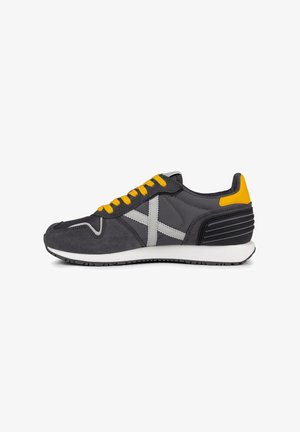 Zapatilla deportiva gris con cordones amarillos, upper de gamuza y malla gris, entresuela blanca, talón negro texturizado y acentos gris contrastantes.