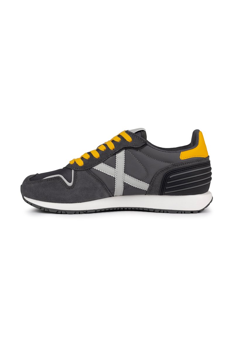 Chaussure de sport grise avec des lacets jaunes, tige en cuir suédé et en maille grise, semelle intermédiaire blanche, talon noir texturé et accents gris contrastants.