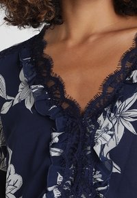 Femme portant un blouse florale bleu marine avec un bord en dentelle froncé le long du décolleté en V profond. Cheveux bouclés visibles à la hauteur des épaules.