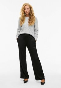 Un pull en tricot gris associé à un pantalon plissé noir taille haute. Le pantalon a une coupe décontractée et des poches latérales, complété par des chaussures noires.
