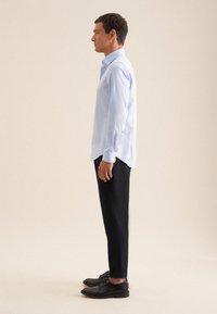 Lichtblauw langemmouwen shirt met knoopmanchetten, gecombineerd met zwarte slim-fit broek en zwarte schoenen, gezien vanuit de zijkant. Neutrale achtergrond.
