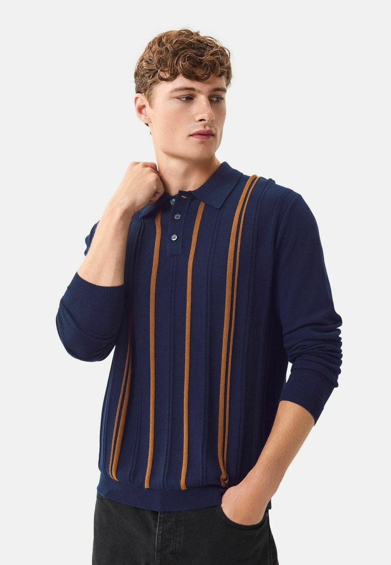 Tumepruun polo-sweater, millel on vertikaalsed oranžid triibud, soonikkoes ja kolme nupuga labi. Pikad varrukad kitsaste mansettidega.