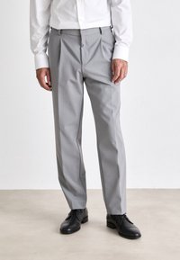 Pantalones gris ajustados hechos de un material suave, con un frente plano, pliegues y bolsillos laterales, combinados con zapatos negros.