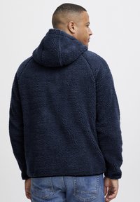 Felpa blu navy in pile con un esterno testurizzato e morbido. Presenta un cappuccio e maniche raglan, abbinata a jeans azzurri.