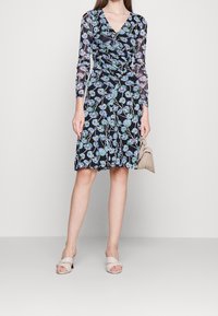 Schwarzes Blumenkleid mit langen transparenten Ärmeln, das blaue Blumen und grüne Akzente zeigt. Das Kleid hat einen V-Ausschnitt und einen ausgestellten Saum. Kombiniert mit hautfarbenen Sandalen.
