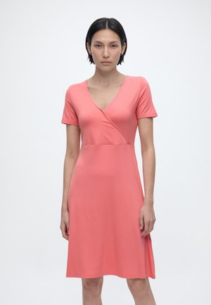 Rövid fekete hajú nő áll, korall rózsaszín rövid ujjú, V-nyakú, térdig érő ruhát visel, egyszínű világos háttér előtt.