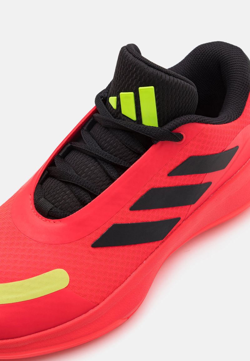 Chaussure de sport rouge avec des accents noirs, tige texturée, détails de logo verts et lacets noirs. Présente un design épuré et une pointe arrondie.