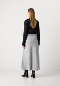 Kvinde med langt, glat hår iført sort bluse, grå ankellang nederdel og sorte hælede ankelstøvler, der står foran en ensfarvet baggrund.